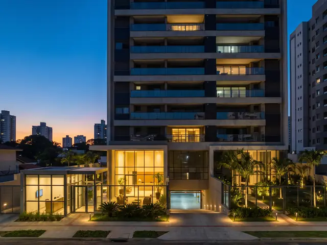 Prédio residencial ao anoitecer, com portaria, garagem e jardim iluminados